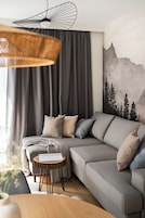 Una sala moderna con un sofá seccional gris, una pequeña mesita de madera y un mural en la pared con un paisaje de montaña.