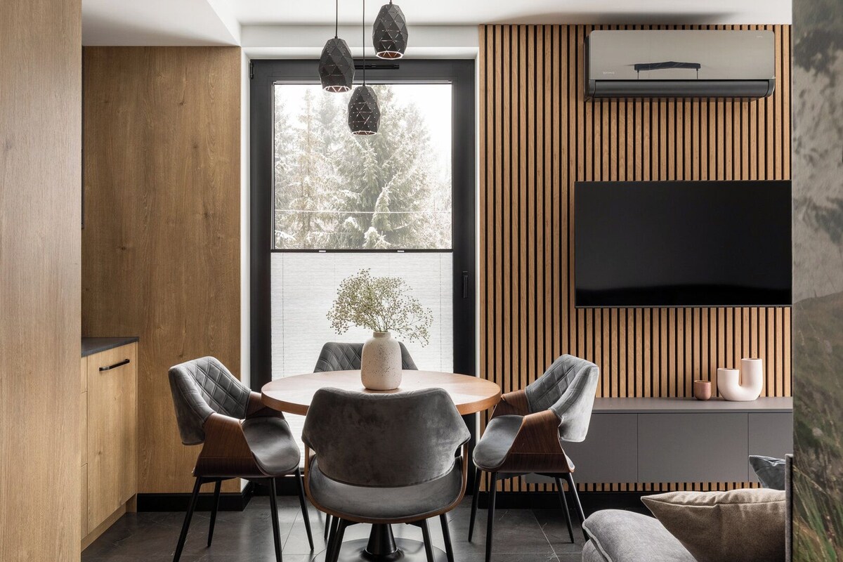 A modern dining area with a round table, grey upholstered chairs, a vase with flowers, and a large window with a view of trees.