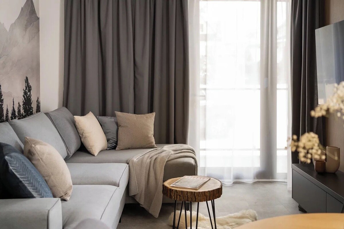 A modern living room with a grey sofa, a round wooden side table, and a large window with curtains.
