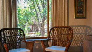 Dos sillones de mimbre con cojines, una mesita redonda con un libro, y vista a zonas verdes a través de una ventana con cortinas.