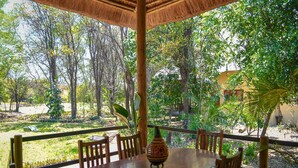 Una terraza de madera al aire libre con una mesa redonda y sillas, rodeada de abundante vegetación y árboles.