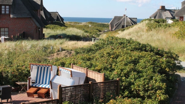 A coastal area with traditional thatched-roof houses, a sandy path, and a beach chair set up for relaxation. - Haus Ruusenhoog - Apartment 7 (Sylt)