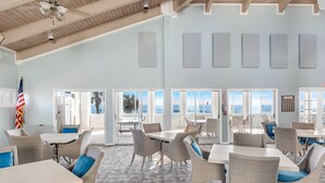 A spacious indoor area with multiple tables and chairs, large windows offering views of the ocean, and a ceiling fan. - Sea Colony Monthly Retreat | Canal View | Steps to Private Beach & Pool | Dog Friendly (Palm Coast)