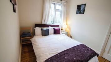 Un lit bien fait, avec une literie blanche et violette, une table de chevet en bois surmontée d’une lampe, et une fenêtre avec des rideaux.