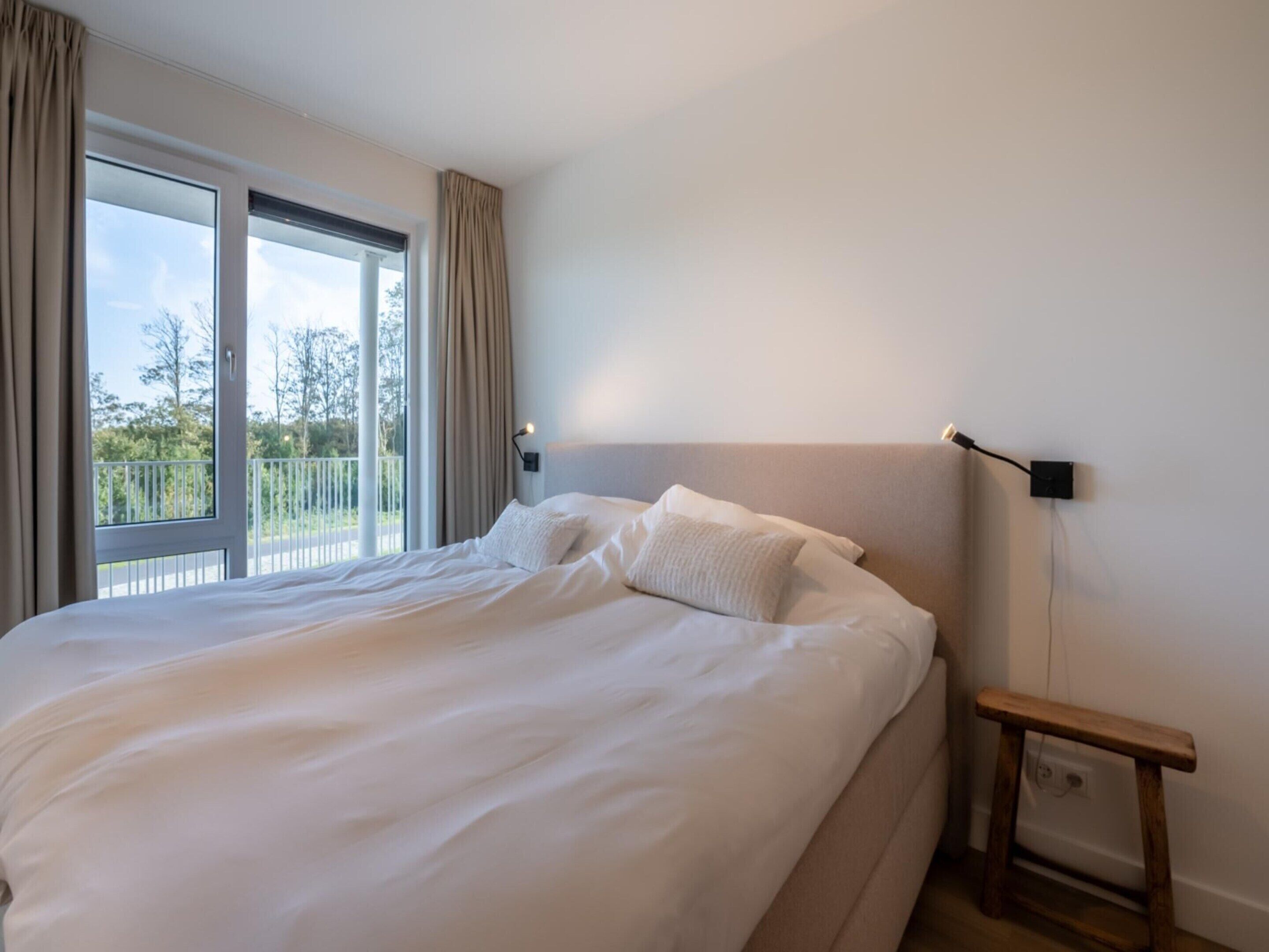 A large bed with white linens, a nightstand with a lamp, and a view of a balcony through a large window.