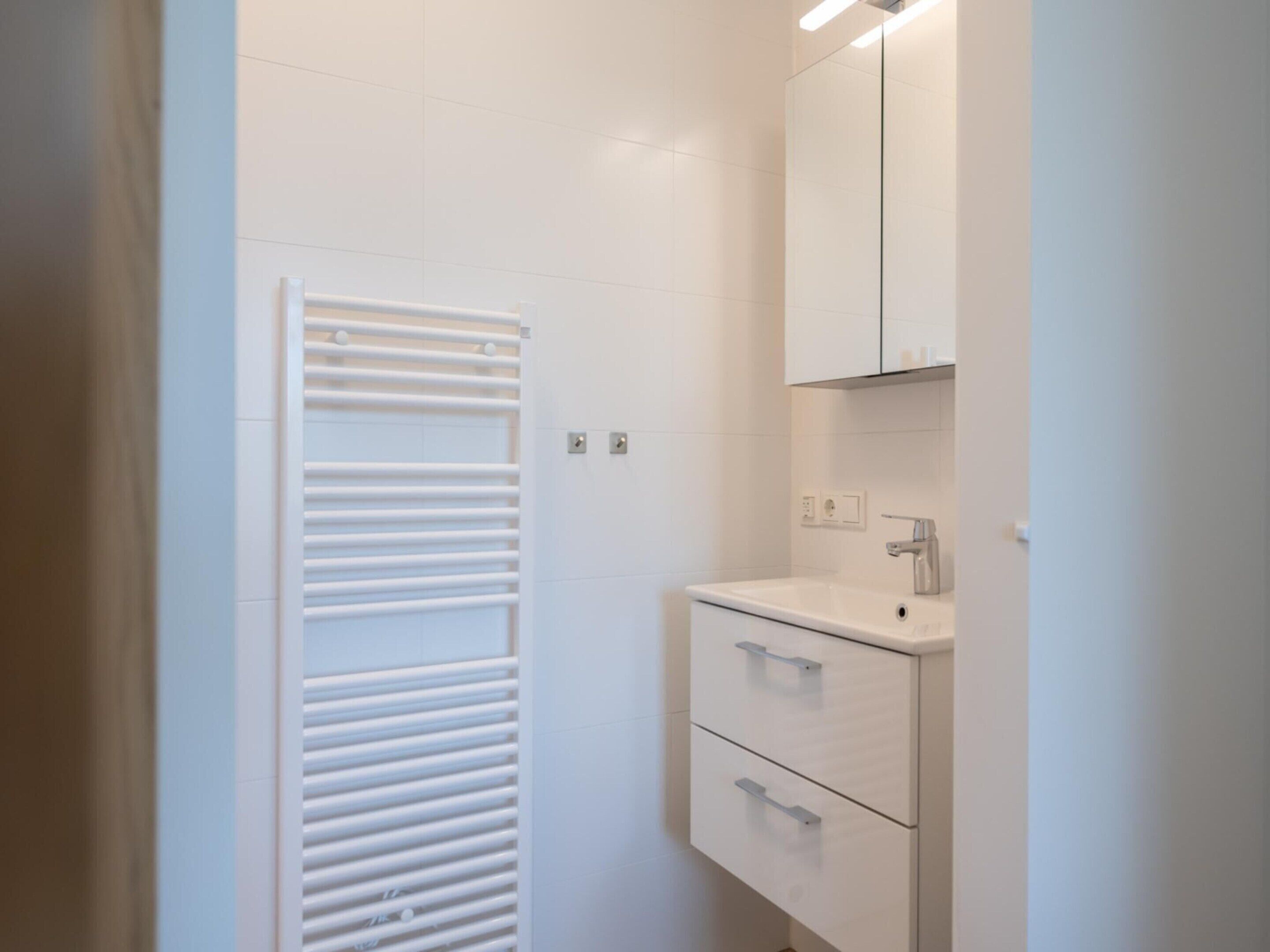 A bathroom with a white tiled wall, a white towel rack, a white sink with a mirror above it, and a white cabinet below.