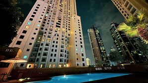 A high-rise hotel with multiple floors, a swimming pool in the foreground, and surrounding tall buildings. - World Class | 1BR | Breathtaking Beach Views (JBR, Dubai)