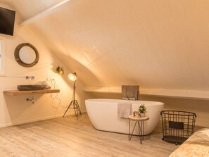 A modern bathroom with a freestanding bathtub, a round mirror, a wooden shelf, a floor lamp, and a small side table with a plant. - Holiday Home Chill on Ameland (Ballum)