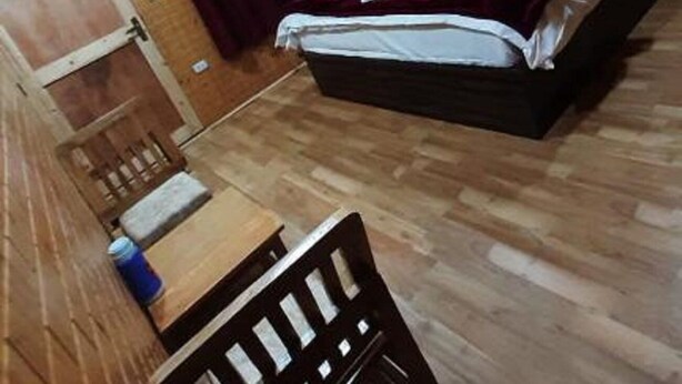 Een kamer met een houten vloer, een bed met wit beddengoed, een houten hoofdbord, een kleine tafel met een blauw voorwerp en een rood gordijn.