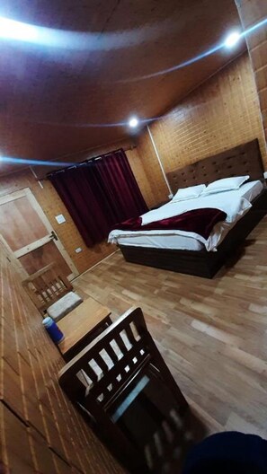 Una habitación con piso de madera, una cama con ropa blanca, cabecera de madera, una mesita con un objeto azul y una cortina roja.