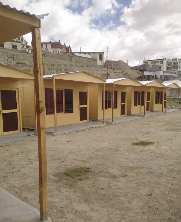 Una fila de edificios amarillos de un piso con ventanas y puertas grandes, ubicados en una zona arenosa con otros edificios y una colina al fondo.
