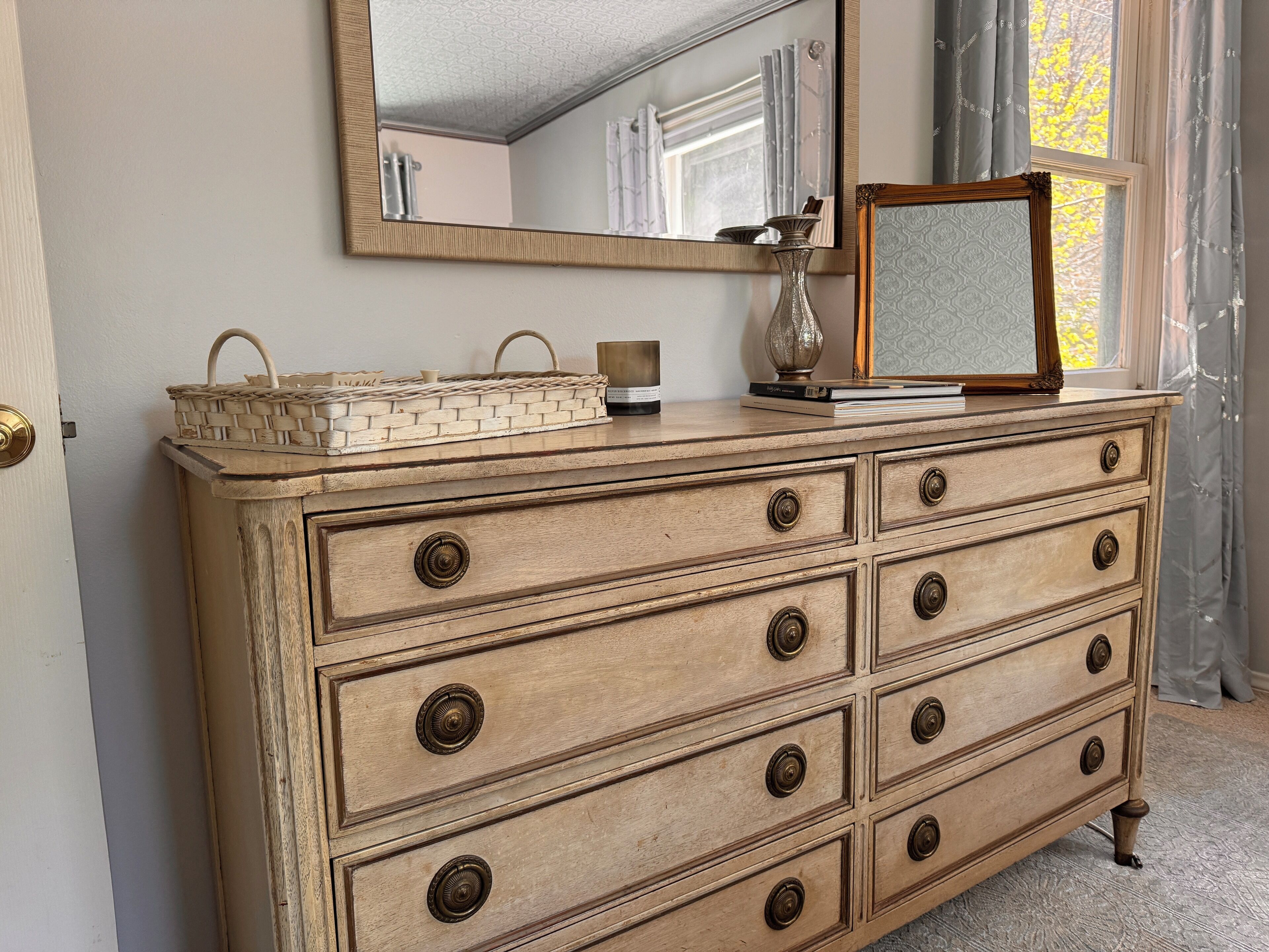 A vintage wooden dresser with multiple drawers and brass knobs, a woven basket, a small vase, and a framed mirror on top.