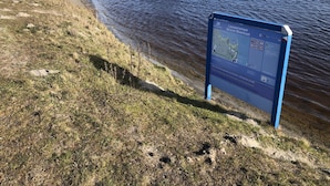 A lakeside area with a blue informational sign, grassy banks, and a row of buildings in the background. - Amazing Holiday Home in Lauwersoog With Sauna (Lauwersoog)
