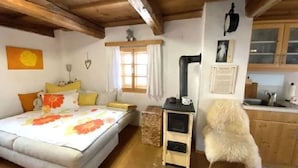 Una stanza accogliente con un letto, un tavolino, una sedia, una finestra con tende, un soffitto in legno e un angolo cottura.