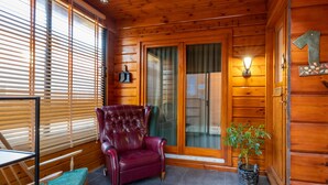 Interior de uma cabana de madeira com uma poltrona de couro, uma cadeira de balanço, uma mesa e um vaso com planta.