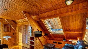 Quarto de sótão aconchegante, com paredes de madeira, claraboia, sofá de couro preto, mesa pequena e televisão embutida.