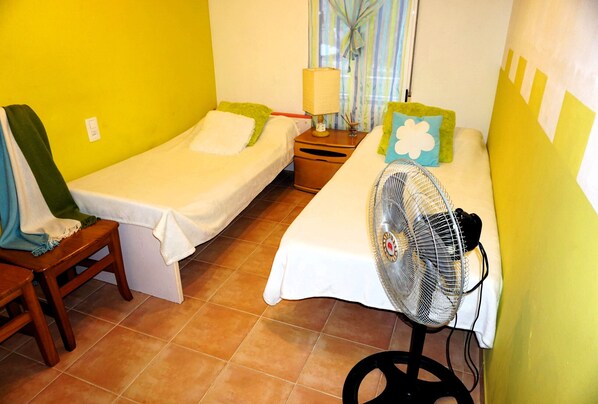A small hotel room with two single beds, a wooden chair, a standing fan, and a nightstand with a lamp.