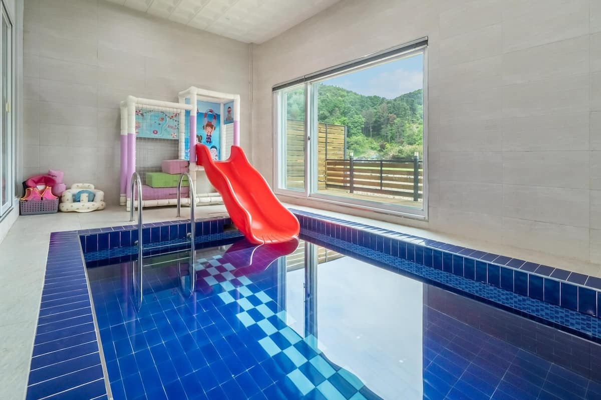 Une piscine intérieure avec un toboggan rouge, un espace de jeux pour enfants et une grande fenêtre donnant sur une zone boisée.