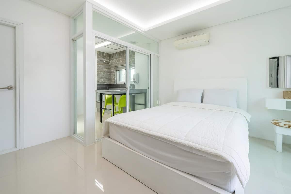 Une chambre moderne avec un grand lit, une petite table de chevet, un miroir et un coin cuisine intégré avec une chaise verte.