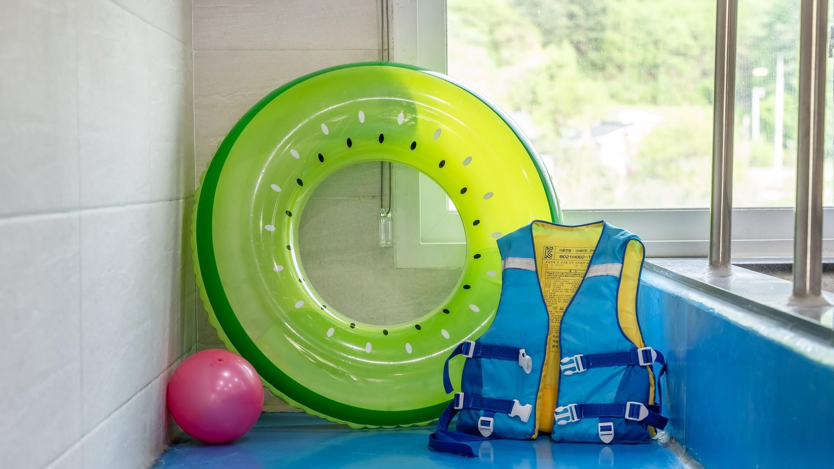 Una ventana con un interior azul, un flotador inflable verde, una pelota rosa y un chaleco salvavidas azul.