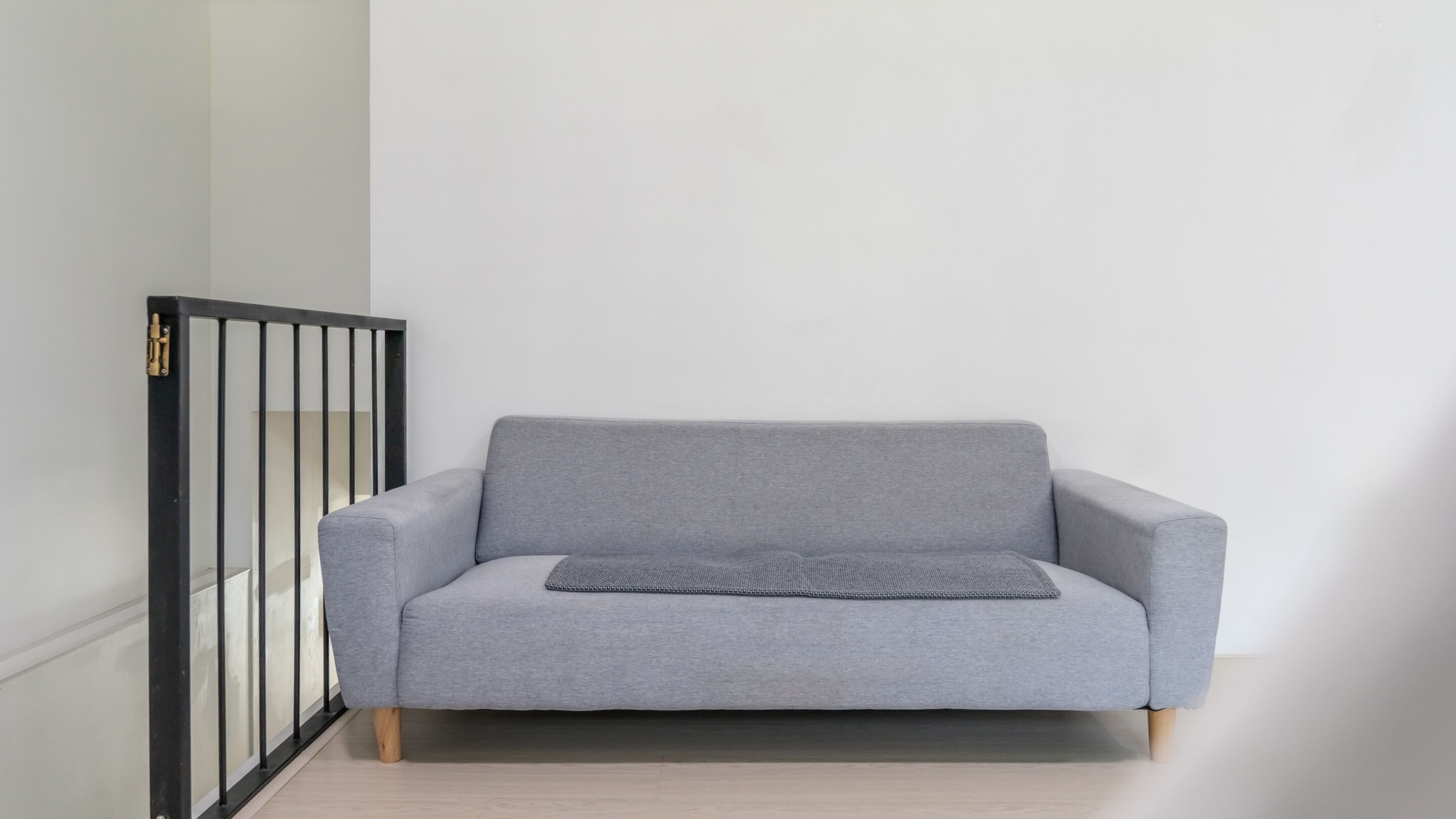 Ein graues Sofa mit Holzbeinen und einem kleinen grauen Kissen, vor einer weißen Wand mit einem schwarzen Metallgitter links.