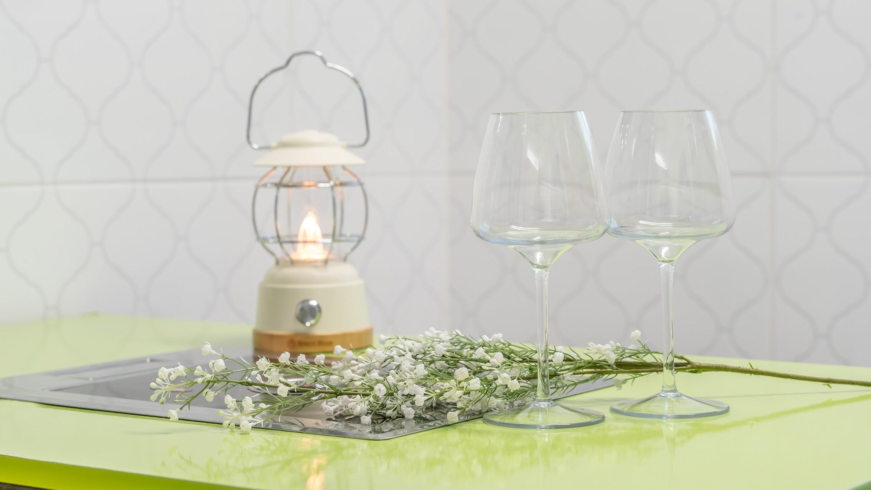 A table with two clear wine glasses, a lit lantern, and a sprig of white flowers on a green surface.