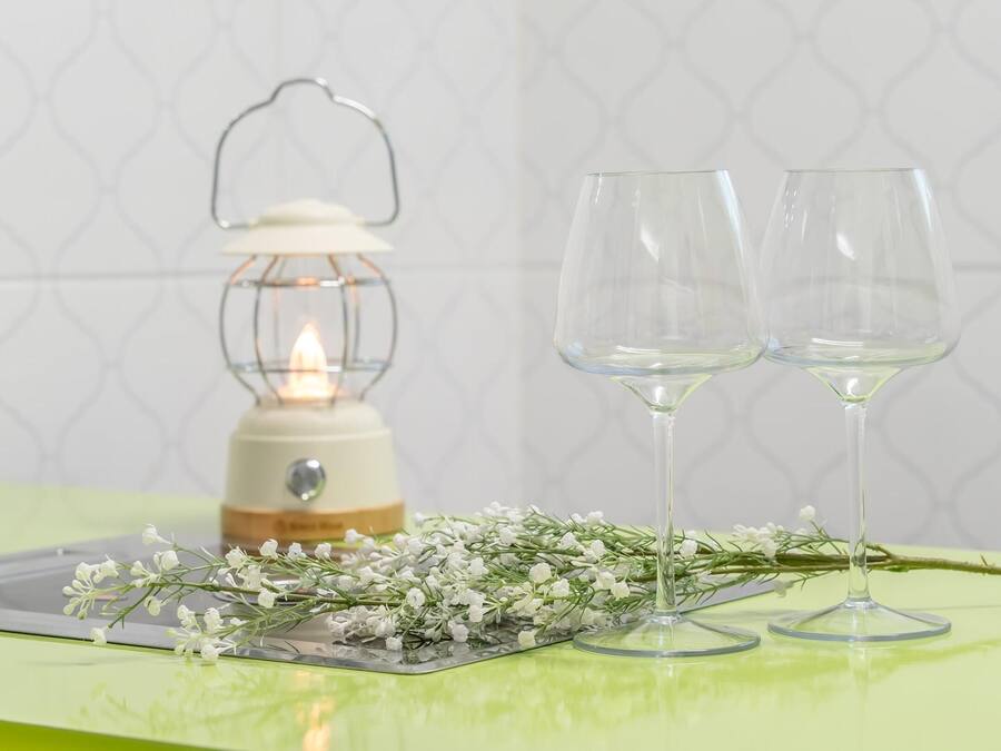 A table with two clear wine glasses, a lit lantern, and a sprig of white flowers on a green surface.