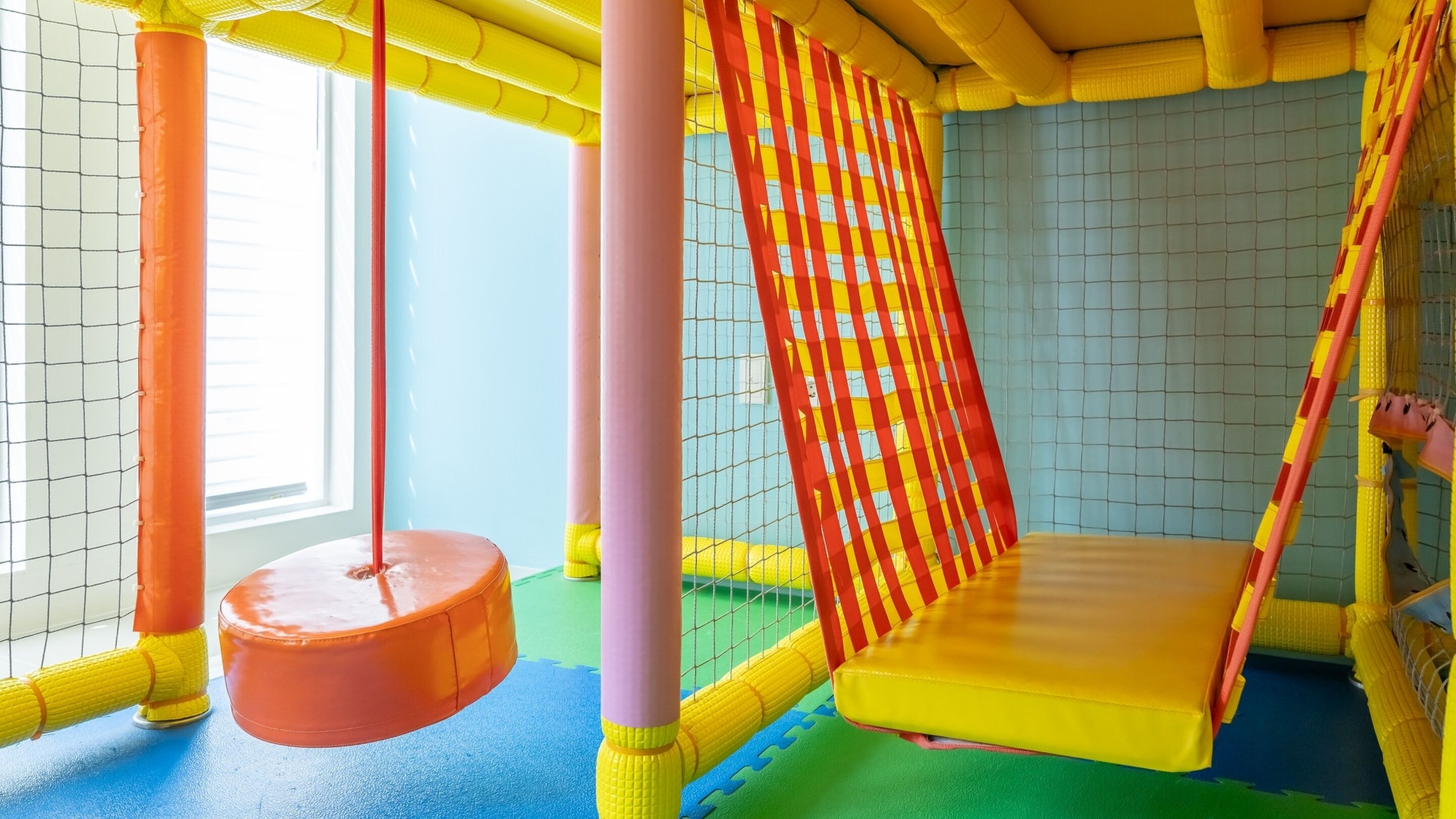 A colorful indoor play area with a swing, padded walls, and a netted ceiling.