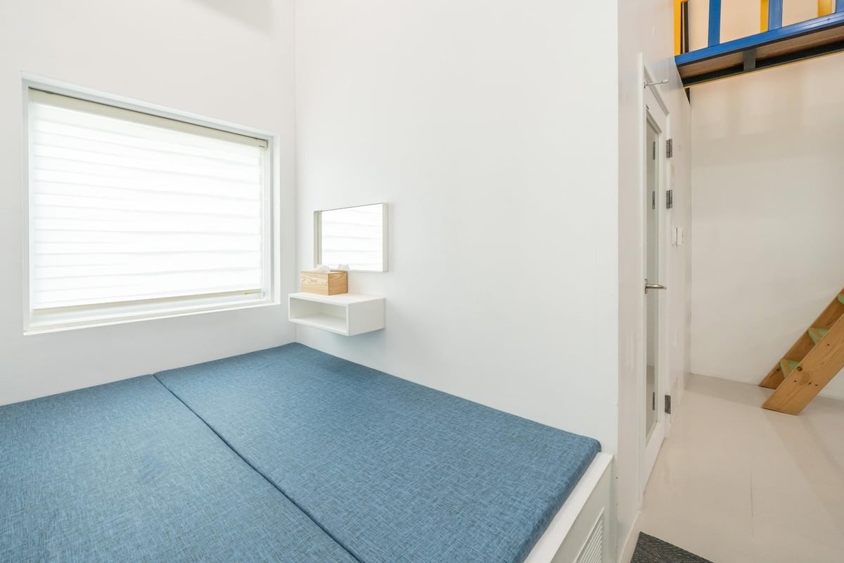 Une pièce avec un matelas bleu, une fenêtre avec des stores, une petite étagère blanche sur laquelle se trouve une boîte, et un escalier en bois.