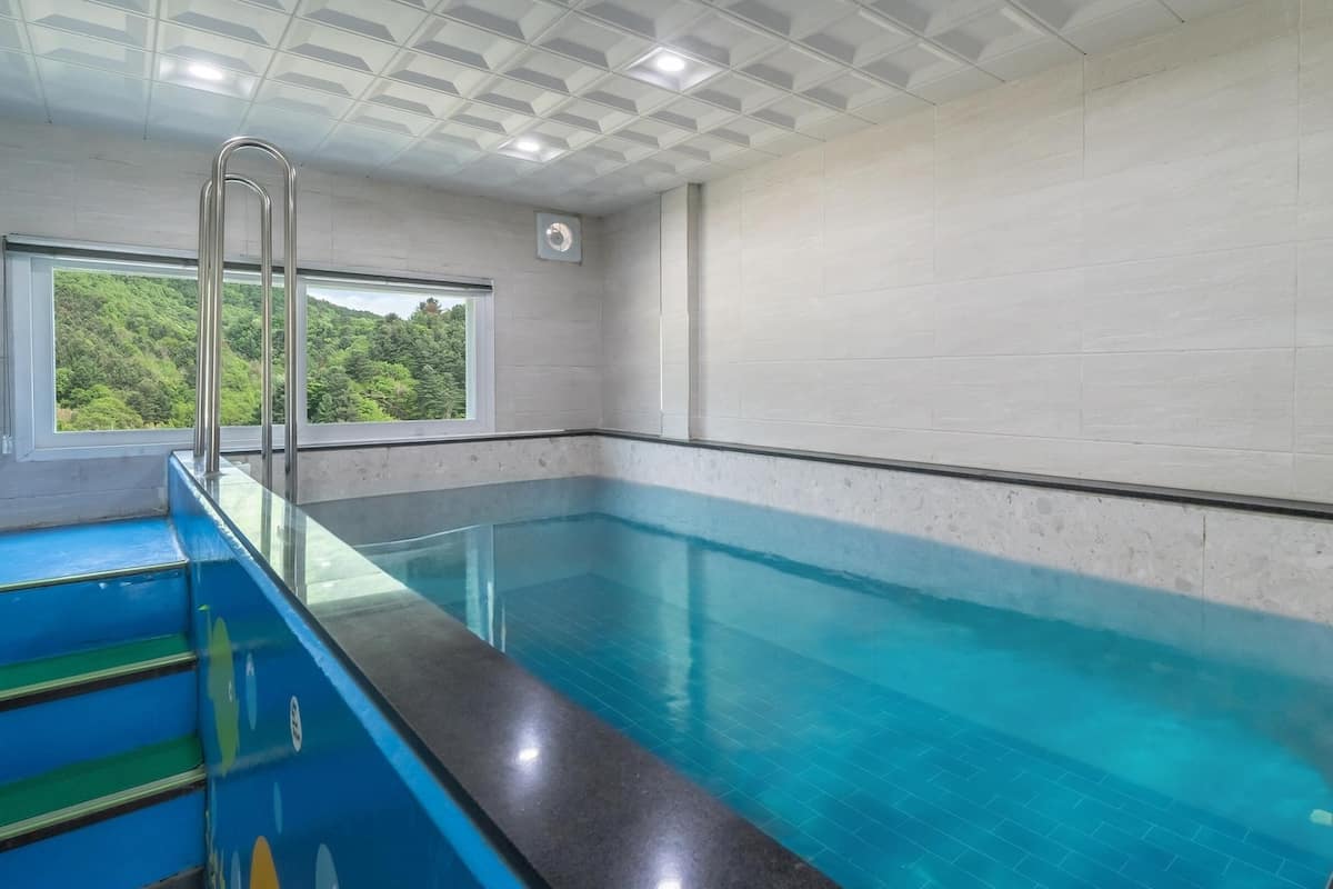 Une piscine intérieure à l’eau d’un bleu cristallin, séparée par une paroi de verre et dotée d’une fenêtre donnant sur un paysage verdoyant.