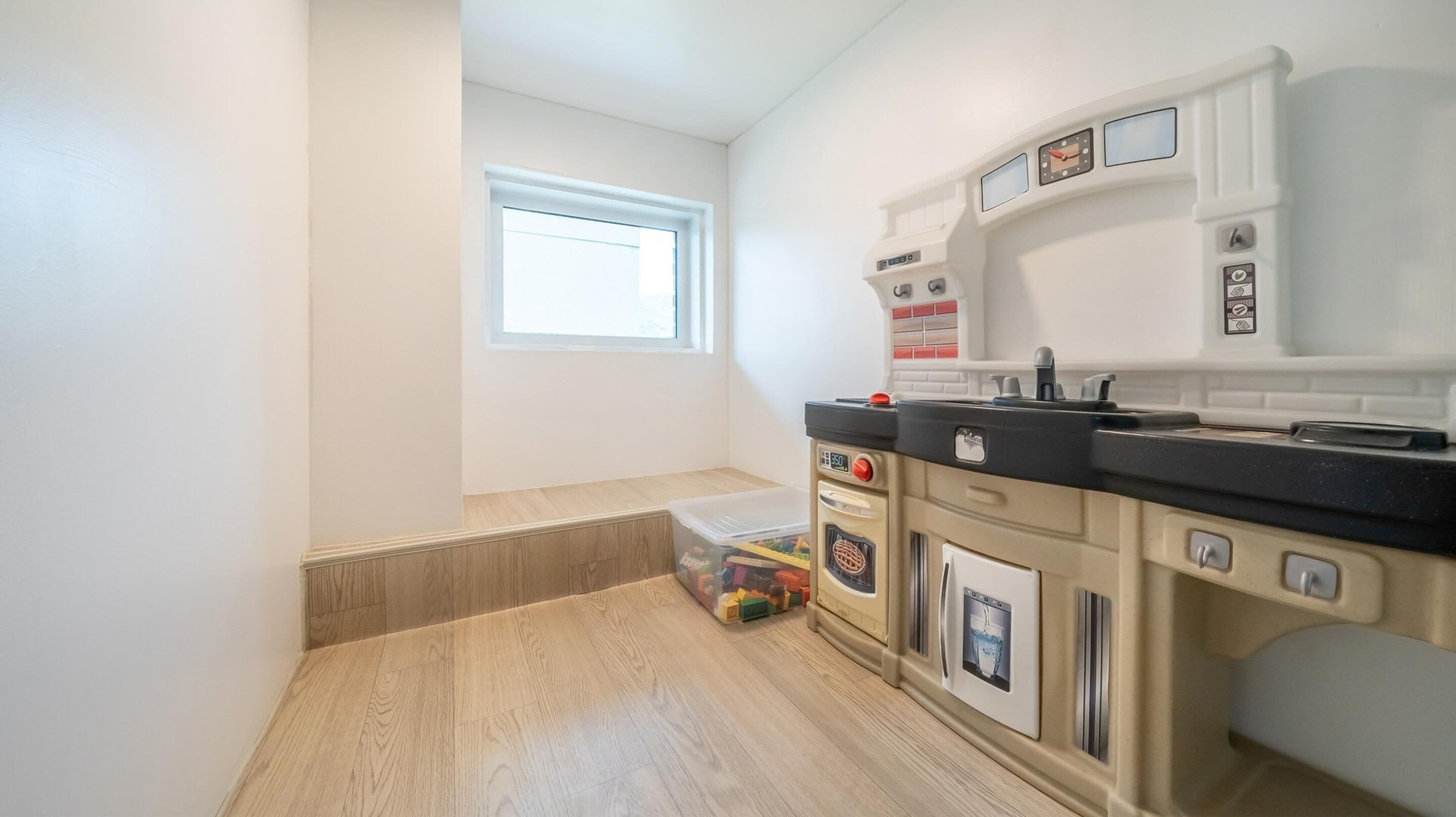 Une cuisine avec une fenêtre, un plancher en bois et un coffre pour ranger les jouets.