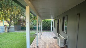 Een overdekt terras met een houten terras, een kleine airconditioning en uitzicht op een gazon met bomen.