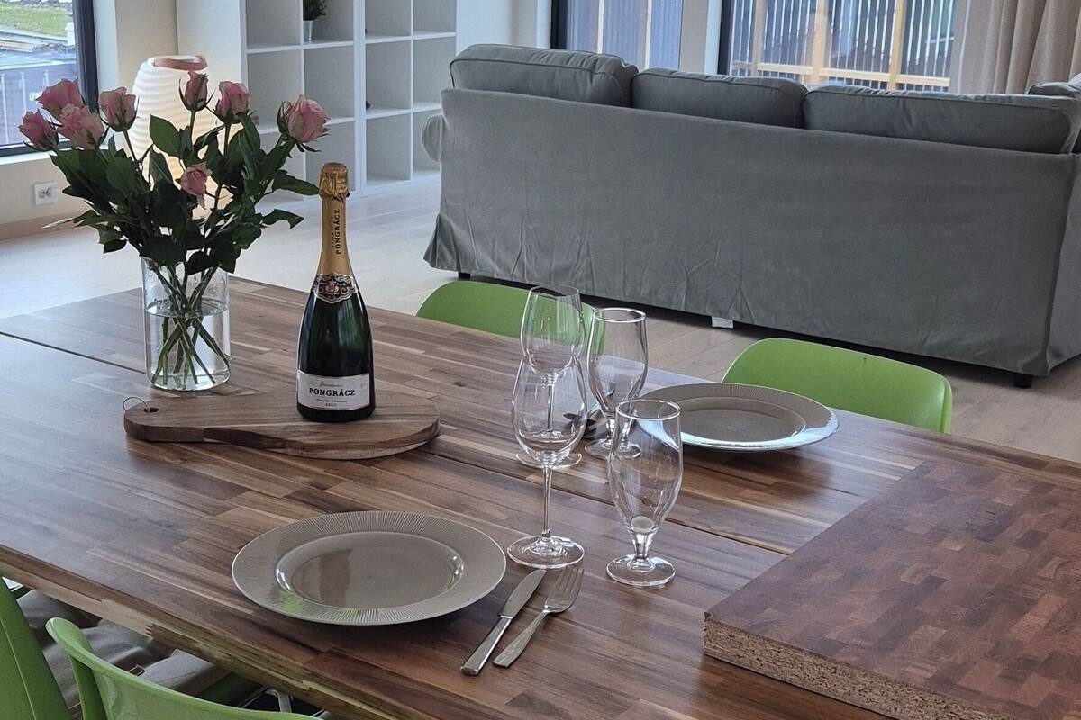 Uma sala de jantar com uma mesa de madeira posta para dois, cadeiras verdes e vista para o mar através de grandes janelas.