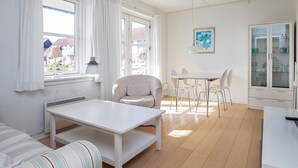 Una habitación luminosa con una mesa blanca, un sofá a rayas y una silla. Hay una ventana con cortinas, una lámpara y un mueble con puertas de cristal.