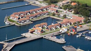 Vista aérea de un puerto deportivo costero con varias embarcaciones amarradas, una serie de edificios con techo rojo y una zona verde con árboles dispersos.
