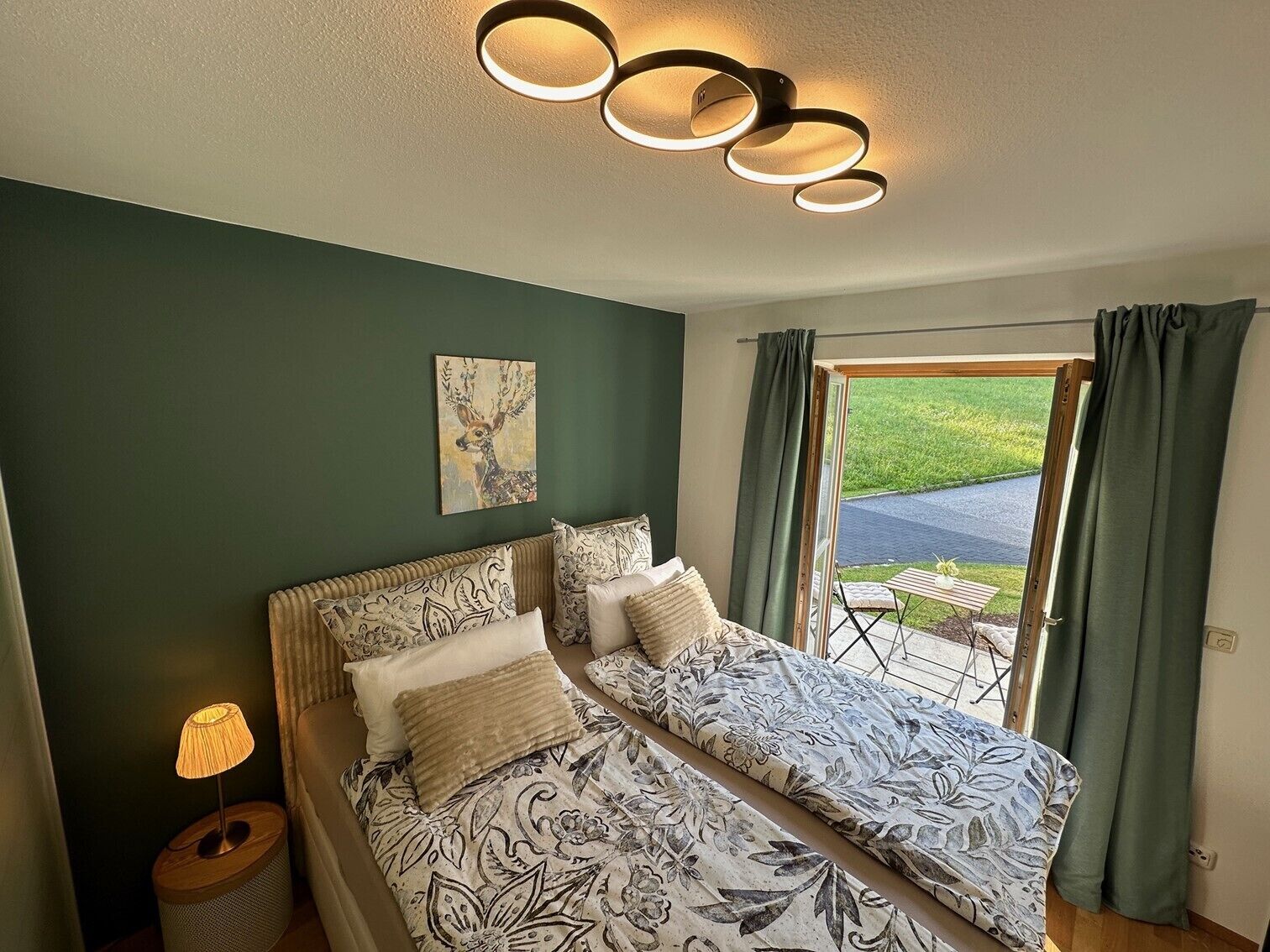 Ein Schlafzimmer mit Bett, Nachttischlampe, Deckenleuchte, einem Gemälde und Blick ins Freie.