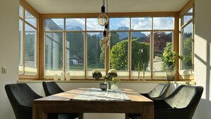 A dining area with a large window, a wooden table, and four chairs. - Ferienwohnung Marianne Bergpanorama (Berchtesgaden)