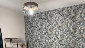 Une chambre à coucher moderne avec un lit, un luminaire suspendu, un mur orné d’un motif de feuilles et une table de chevet sur laquelle se trouve une tasse.