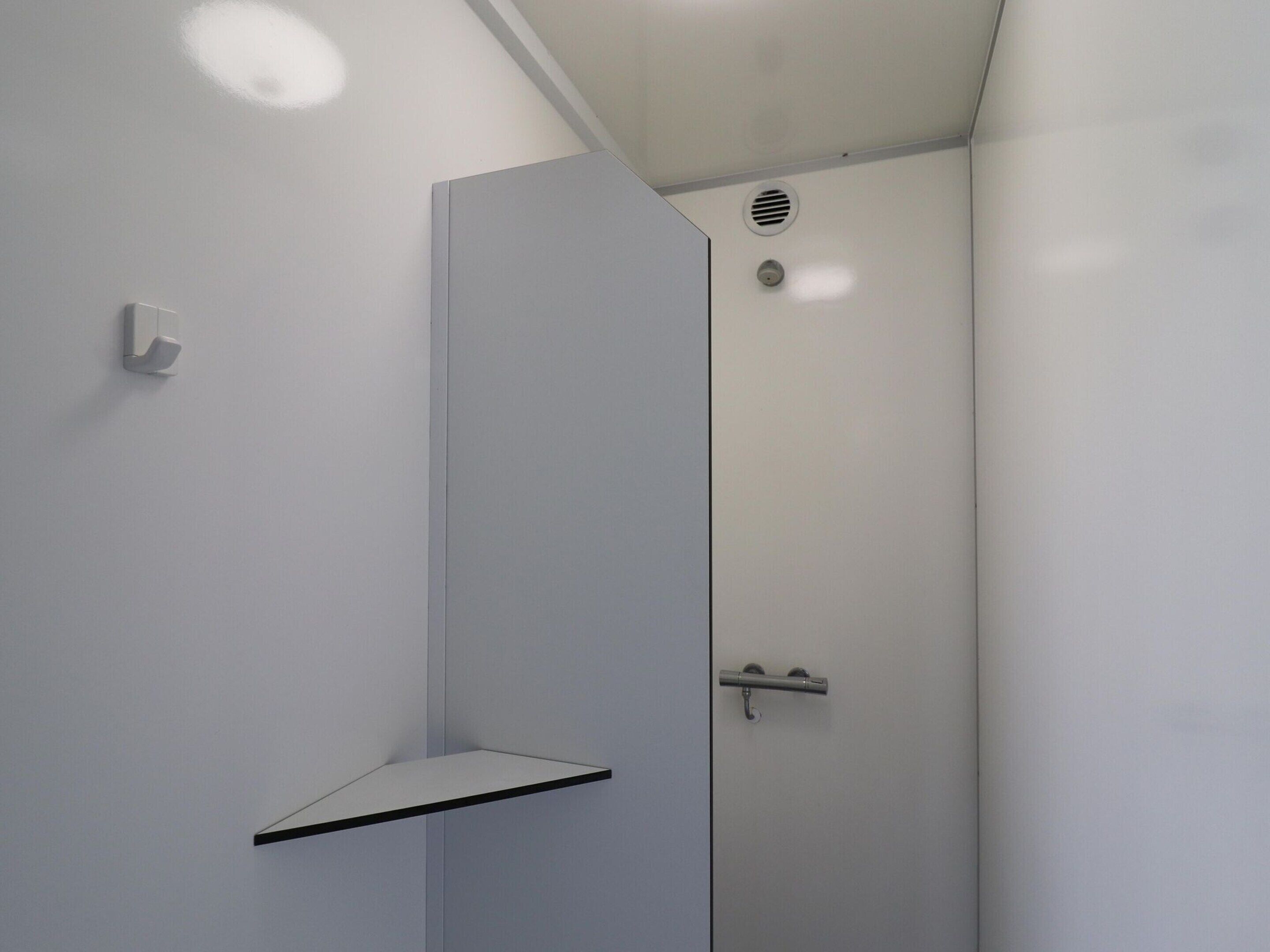 A modern bathroom with a white wall, a corner shelf, a door with a handle, and a vent on the ceiling.