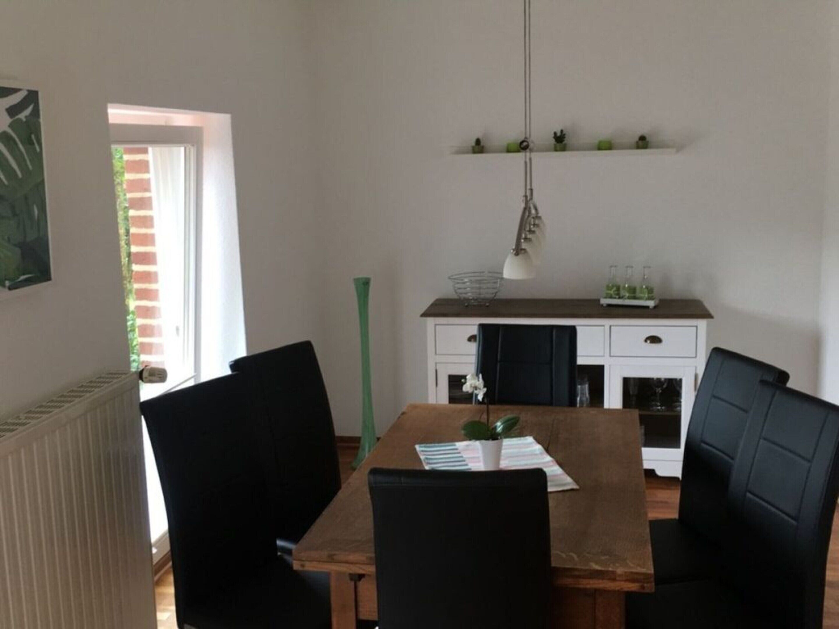 A dining area with a wooden table, black chairs, a hanging lamp, a small potted plant, and a white cabinet with shelves above.