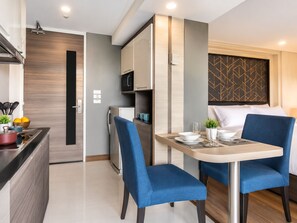 A compact hotel room with a kitchenette, a dining area with two chairs, a bed with a headboard, and a small potted plant.