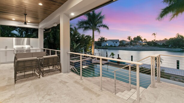 A modern outdoor kitchen with a pool view, a dining area with wicker furniture, and a staircase leading to a balcony with a railing overlooking a body of water. - Relax with Pool, Hot Tub, Fire Pit & Waterfront Views (Marco Island)