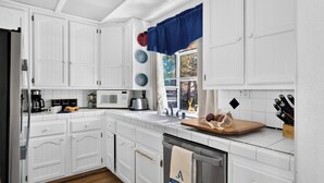 Una cocina con armarios blancos, microondas, cafetera y ventana con cortina azul.