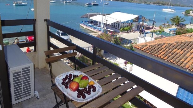 Un balcone con un tavolo su cui è posata una porzione di frutta, vista su una marina con barche e un edificio con tetto bianco.