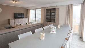 A modern living room with a grey sofa, a dining table with chairs, a TV, and a cabinet with glassware. - Spacious Coastal Retreat (Middelkerke)
