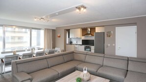 A modern living room with a sectional sofa, a wooden coffee table with decorative items, and a kitchen area with a refrigerator and oven. - Spacious Coastal Retreat (Middelkerke)