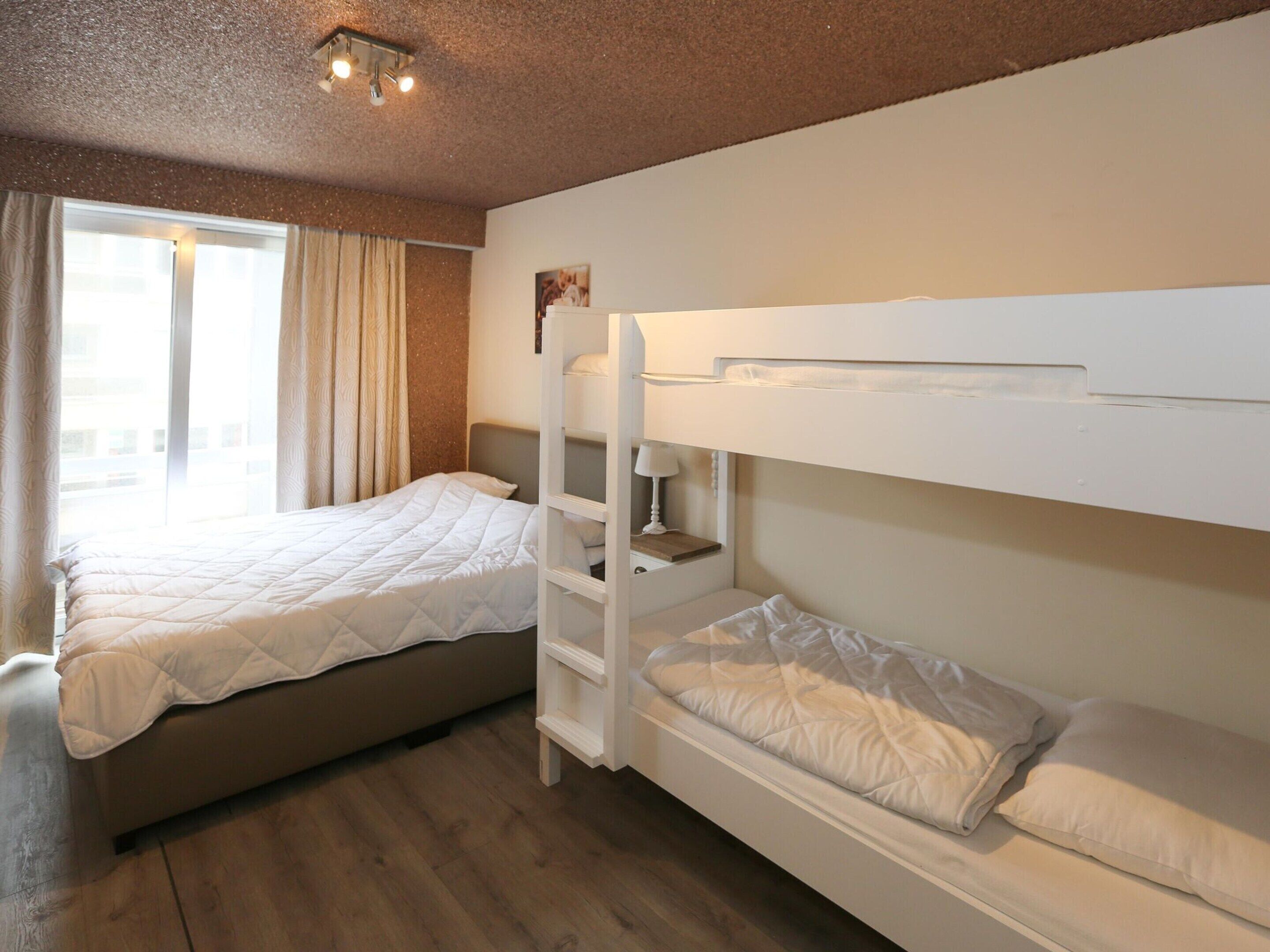 A room with two beds, one with a bunk bed and the other a single bed, wooden flooring, a window with curtains, and a ceiling light fixture.