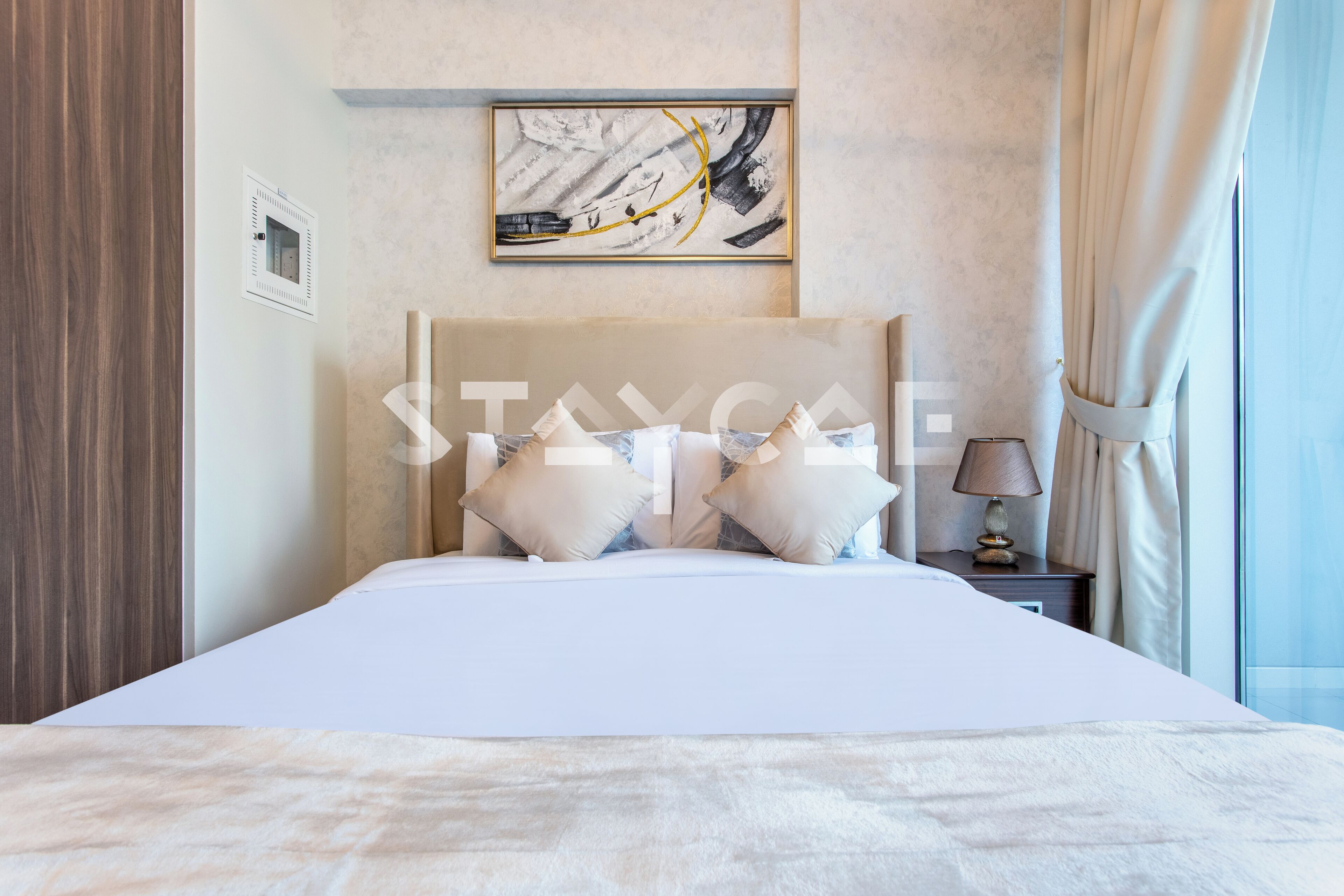 A modern hotel room with a large bed, a headboard with a framed artwork, a bedside table with a lamp, and a view of the outdoors through a window with curtains.