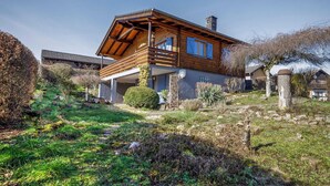 Une maison en bois dotée d’un balcon, entourée d’un jardin où poussent différentes espèces de plantes, et agrémentée d’une structure en pierre.