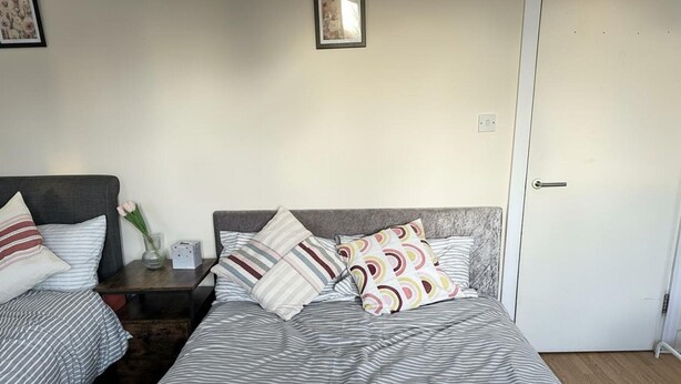 Uma cama de casal com roupa de cama listrada e várias almofadas, uma mesa de cabeceira com um vaso e uma luminária, e uma porta à direita.